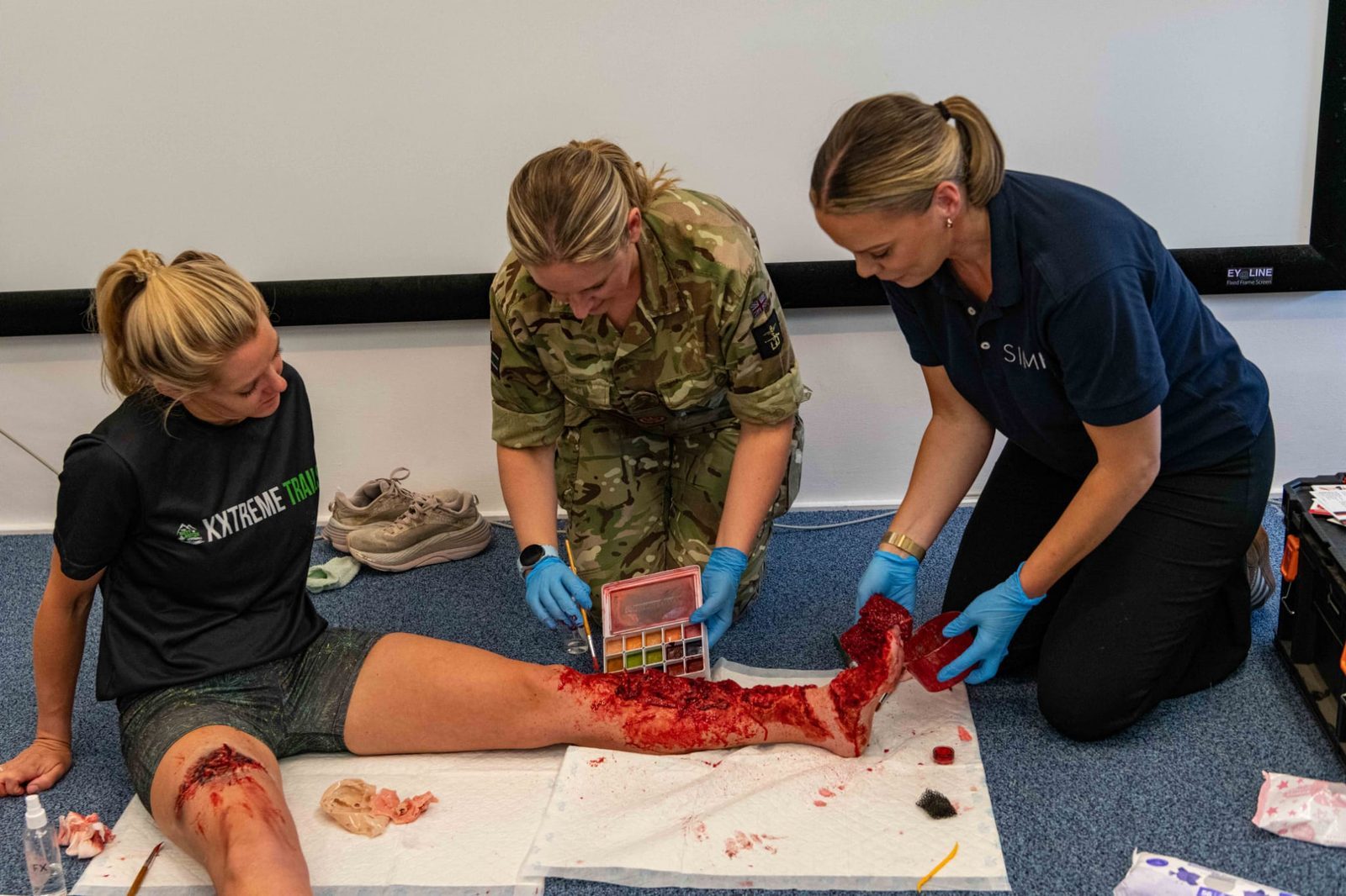 Three people working together on leg moulage during a training session, two applying with brushes and palette while the third sits as the casualty