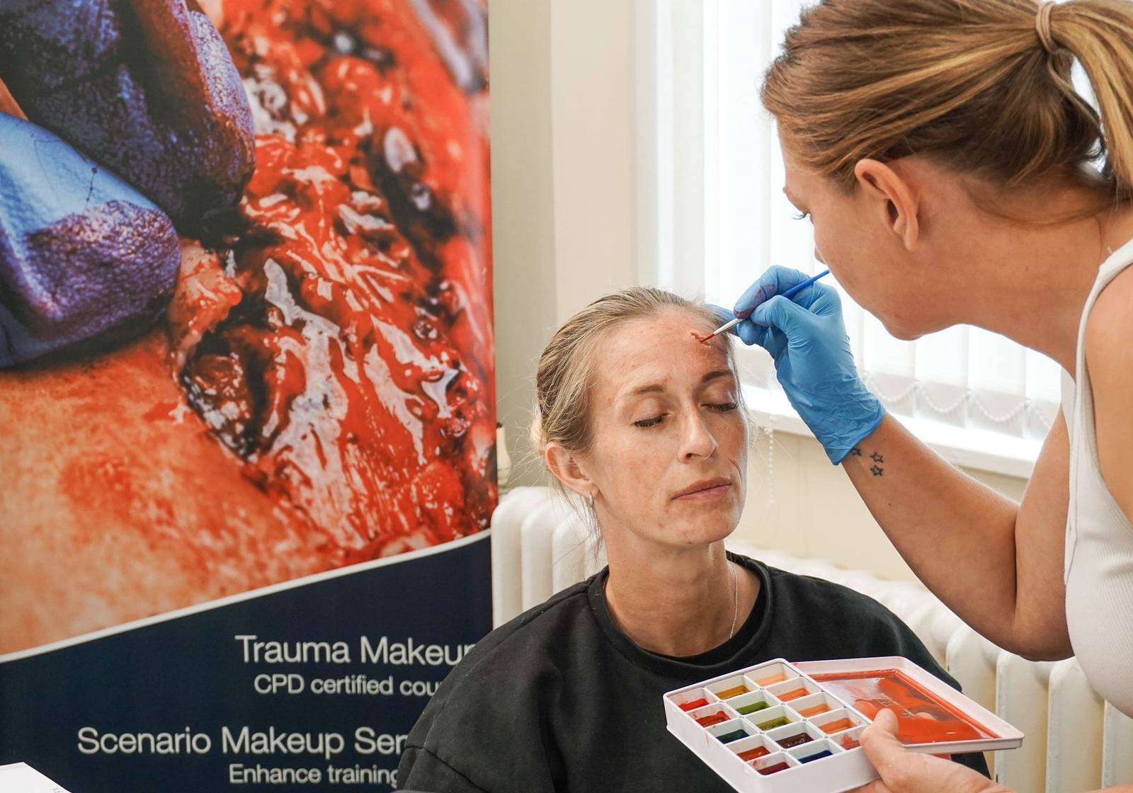 A trainer applying facial moulage to a seated delegate during a CPD certified course, the SIMFX course banner behind them