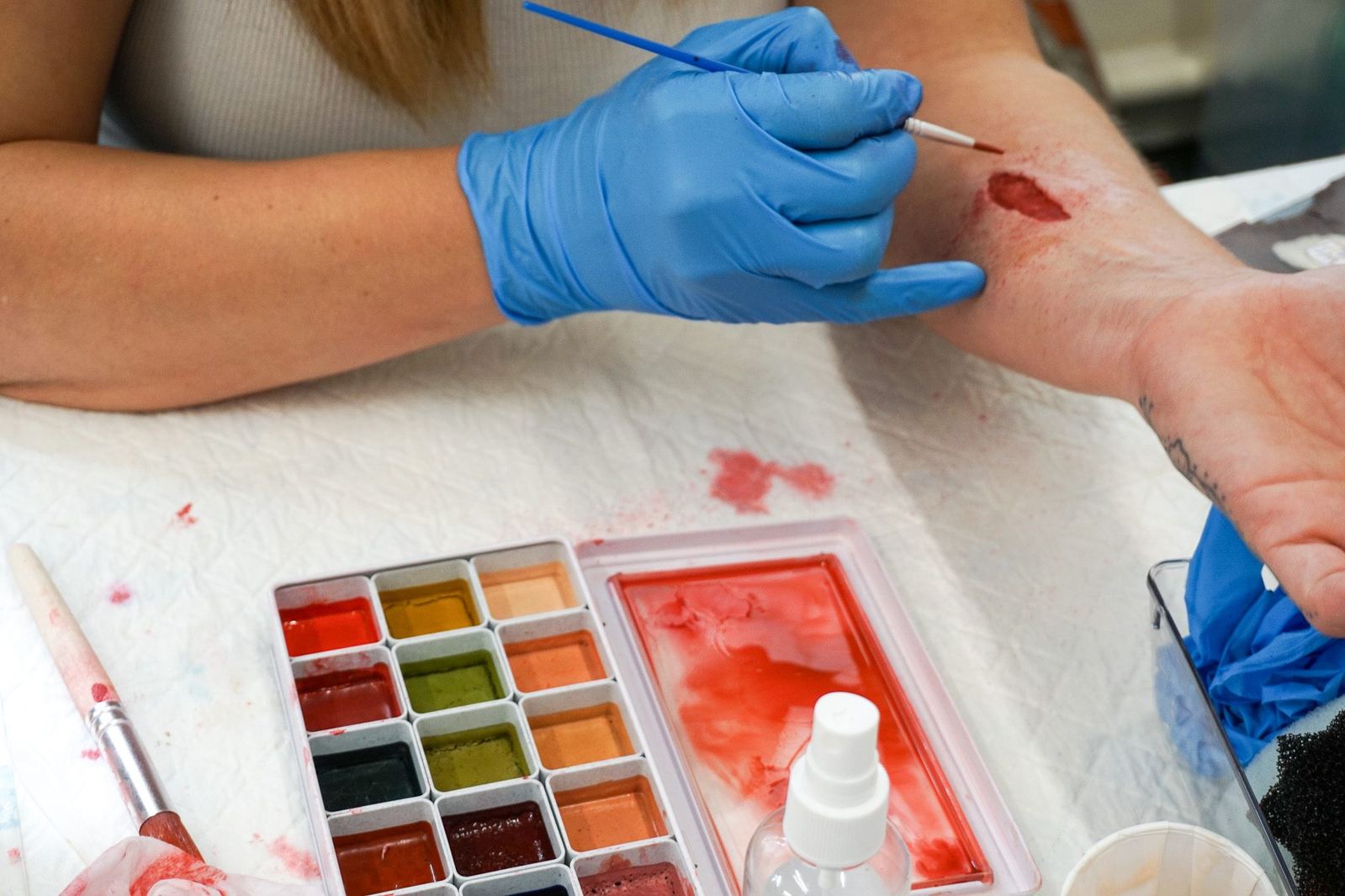 Gloved hands painting colour into a forearm wound over a bench towel, palette in frame